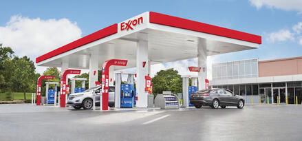 An Exxon station on a sunny day with clear blue skies. Two cars are at the station fueling up.