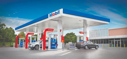 A Mobil station on a sunny day with clear blue skies. Two cars are at the station fueling up.