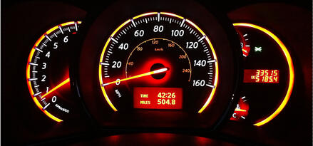 A close-up of a vehicle dashboard at night showing the speedometer, fuel gauge, and temperature gauge. There are bright red and yellow lights illuminating the guages.