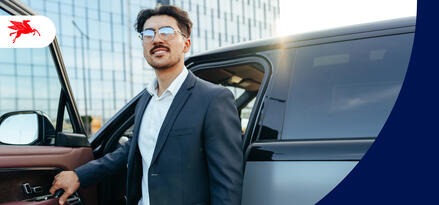A young man in a suit jacket with glasses and a mustache is getting out of his SUV in front of a glass office building.