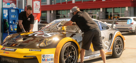 A black and yellow Porsche race car outside of a Mobil branded station with two men fueling and cleaning it.