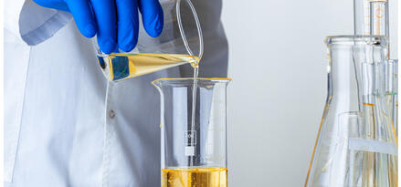 A gloved hand pouring liquid from one beaker to another in a lab setting.