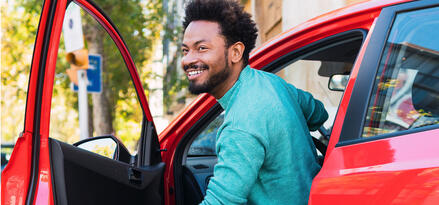 A man with black hair and a turquoise shirt is getting out of his red SUV.