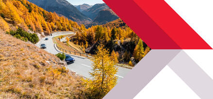 Several blue cars are driving up a winding road through mountains covered with fall-colored trees.