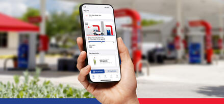 A hand is holding a phone with the screen featuring the Exxon Mobil Rewards+ app, in front of an Exxon station.