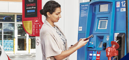A brunette woman at a Mobil gas pump, holding her phone in her hands to access the Exxon Mobil Rewards+ app