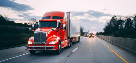 A semi truck with a red cab and white intermodal container driving down a semi-busy highway at dusk. There is another semi truck and car in the distance.