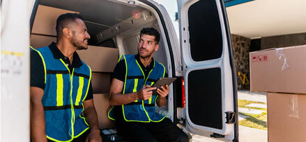 Two men in collared shirts with blue and neon yellow striped vests leaning on the back of a van. The van's back doors are open with visible packages in the vehicle. One man is holding an iPad.