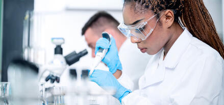 A man and woman in white coats with blue protective gloves in a lab. The man is looking in a microscope and the woman is wearing goggles and testing fuel.