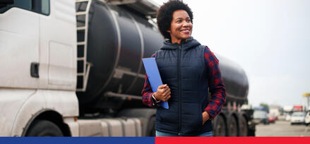 A smiling woman in a puffer vest holding a blue folder and standing beside a truck that transports fuel