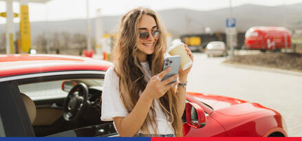 A young woman in sunglasses holding a coffee and smiling at her phone as she stands against her red car on a sunny day.