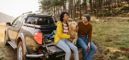 A man and woman sitting on the back of a pickup truck laughing and smiling with a golden retriever in between them.