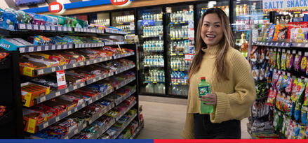 A woman smiling and holding a soda in the candy aisle of an ExxonMobil branded convenience store.
