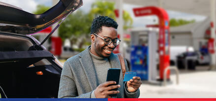 A mean wearing a turtleneck sweater, blazer, and glasses is standing near his car at the gas station looking at the back of his Exxon Mobil Smart Card+ credit card with his phone in his other hand.