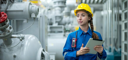 A woman in a blue jumpsuit and yellow hard hat holding an iPad near industrial-looking storage tanks, signaling she is conducting a fuel quality check.