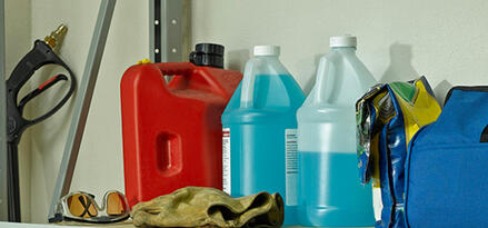 A red gasoline container, two bottles of wiper fluid, and safety items, including plastic goggles and gloves, on a garage storage shelf