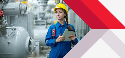A woman in a blue jumpsuit and yellow hard hat holding an iPad near industrial-looking storage tanks, signaling she is conducting a fuel quality check.