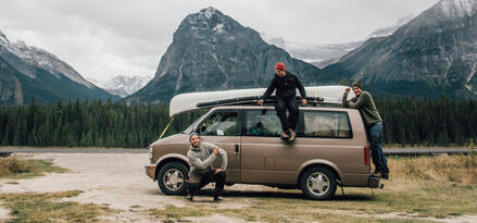 Three men standing on or around a brown van with a canoe strapped to the top in a mountainous area.