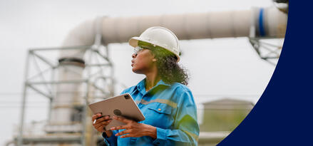A fuel quality surveyor on-site wearing a hard hat and safety goggles, holding a tablet and looking into the distance.