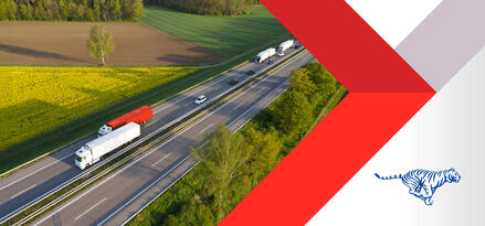 Two semi trucks driving down a quiet highway next with a field on on side and trees on the other.